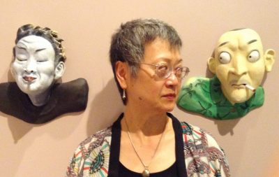 Margo Machida at the Honolulu Museum of Art, standing between two sculptures of faces, mounted on the wall.