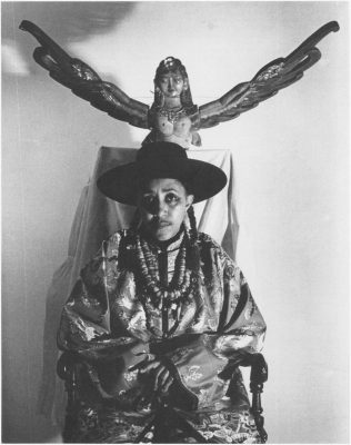 Photo of Camille Billops sitting in a chair, wearing a wide-brimmed hat, under a sculpture of a woman with wings.