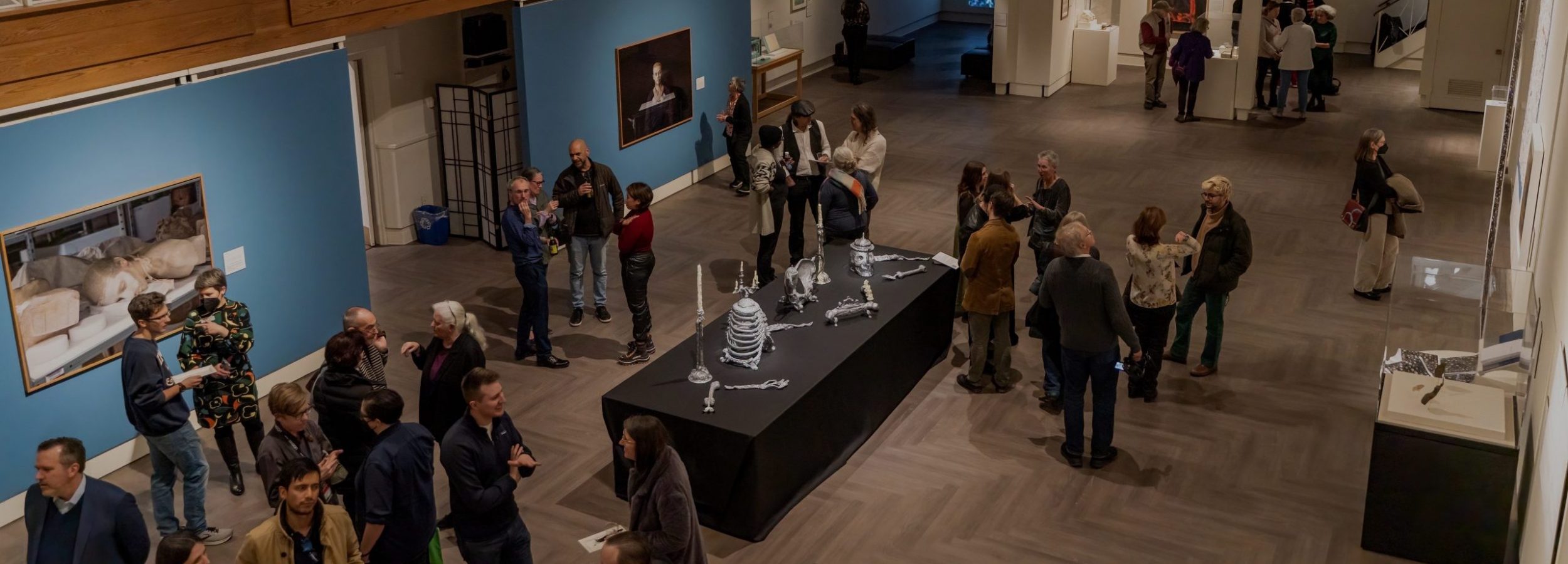 An aerial view of people gathered at the Seeing Truth exhibition at the Benton Museum. Art is hanging on blue walls and in the middle of the room a table covered in a black table cloth holds silver objects.